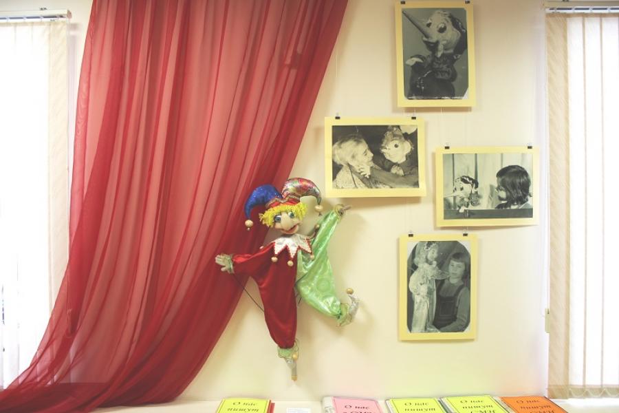 Изображен фрагмент экспозиции, с тканью символизирующей театральный занавес, перед ней яркая театральная тростевая кукла Петрушка. Рядом на стене четыре фотографии, рассказывающие об истории Подпорожского театра кукол.